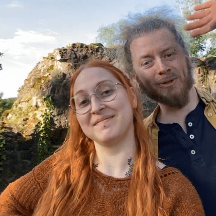 Das Bild zeigt zwei Personen vor einer natürlichen Felsformation. Die Person links hat langes, rotes Haar und trägt eine Brille sowie ein braunes Oberteil. Sie lächelt in die Kamera. Die Person rechts hat graues, lockiges Haar und einen Bart. Sie trägt ein dunkelblaues Hemd mit einem braunen Hemd darüber. Der Himmel im Hintergrund ist klar und blau. Es sieht nach einem sonnigen Tag in einer grünen, natürlichen Umgebung aus.